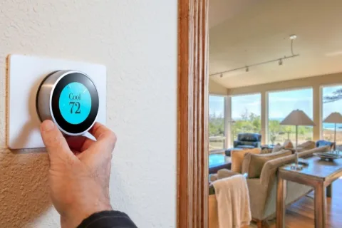 Hand adjusting smart thermostat set at 72 degrees in modern living room with large windows and natural light