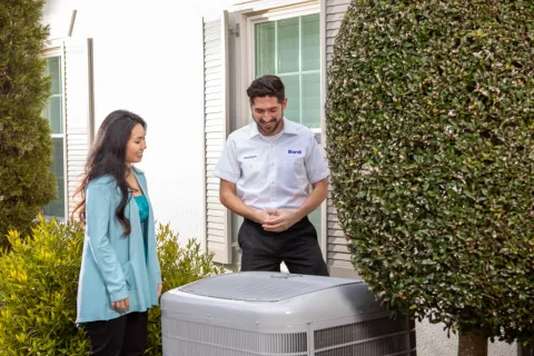Technician explaining HVAC unit to woman outside house near trimmed tree and bushes on sunny day