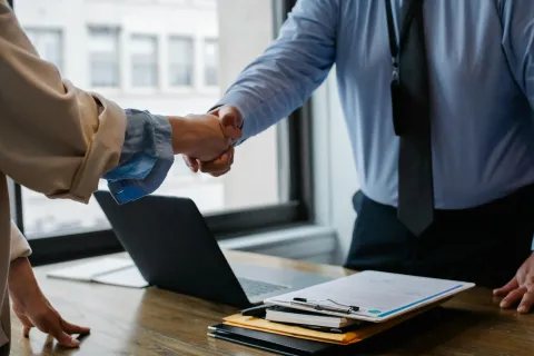 Two professionals shaking hands over a desk with a laptop and documents in a bright office setting