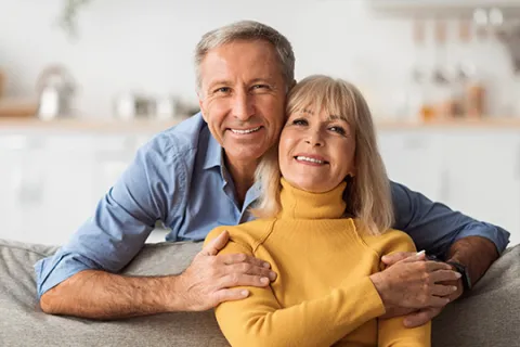 Smiling mature couple hugging on a couch in a cozy, modern living room with soft natural light.