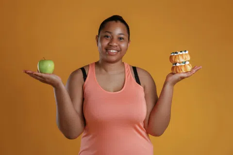 Smiling woman in coral tank top holding a green apple in one hand and stacked cream-filled pastries in the other.
