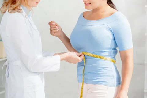 Healthcare professional measuring woman's waist with yellow tape measure in a medical setting.