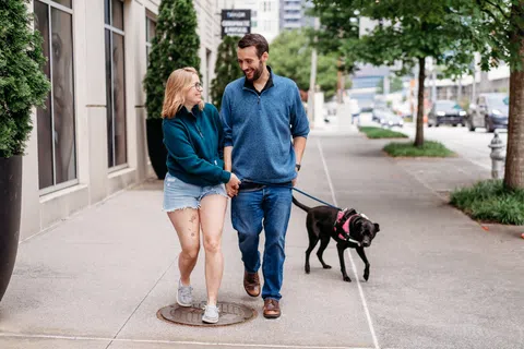 A couple walks their dog hand in hand along a tree-lined city sidewalk, illustrating the everyday outings people plan around after weight loss surgery.