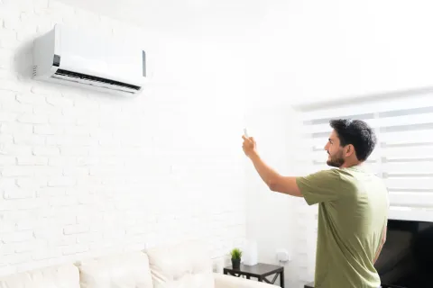 Man in green shirt using remote to control wall-mounted air conditioner in a bright modern living room.