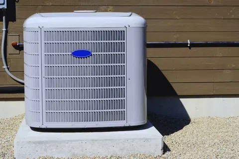Outdoor central air conditioning unit installed on a concrete pad beside a wooden house exterior wall.