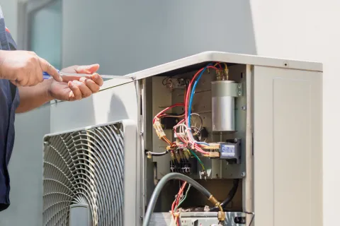 Technician repairing an air conditioning unit with exposed wires using a screwdriver.