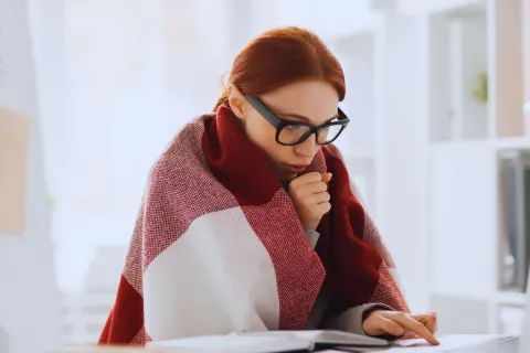 Woman wrapped in a blanket working on a laptop in a bright home office feeling cold.