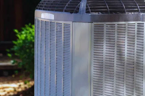Outdoor central air conditioning unit with metal casing and ventilation grilles in a backyard setting.
