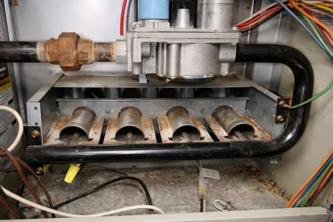 Rusty furnace burners and pipes inside a gas heating system with connected wires and metal components.