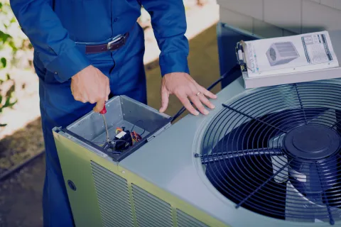 Technician in blue uniform repairing an outdoor air conditioning unit with a screwdriver and manual nearby