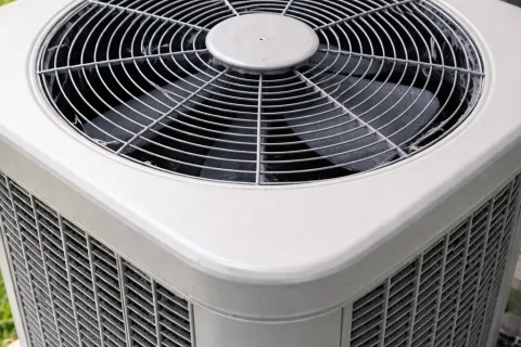 Outdoor home air conditioning unit with metal fan and protective grille installed near a house wall.