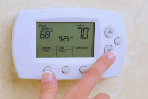 Close-up of hands adjusting a digital thermostat set to 68 degrees with indoor temperature of 70 degrees.