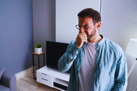 Man with glasses sneezing or coughing in modern living room with TV and lamp in background