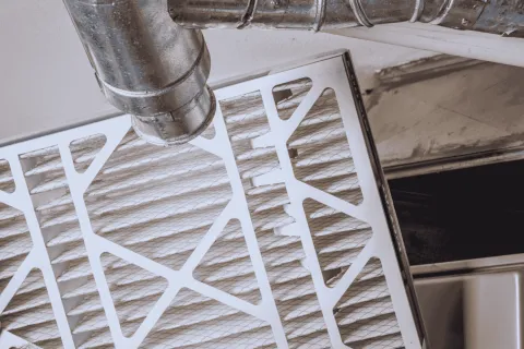Hand holding a clean white air filter near metal ducts inside a furnace room for HVAC system maintenance
