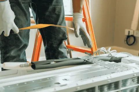 Technician wearing gloves measuring parts during air conditioner installation with tools and ladder nearby