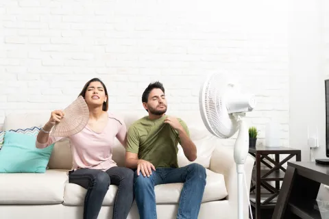 Young man and woman sitting on sofa using fan and hand fan to cool off in hot room.