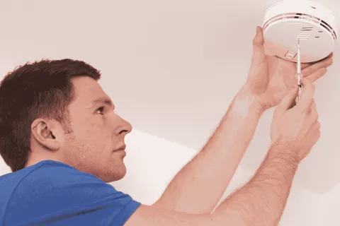 Man in blue shirt installing or repairing a ceiling smoke detector with a screwdriver indoors
