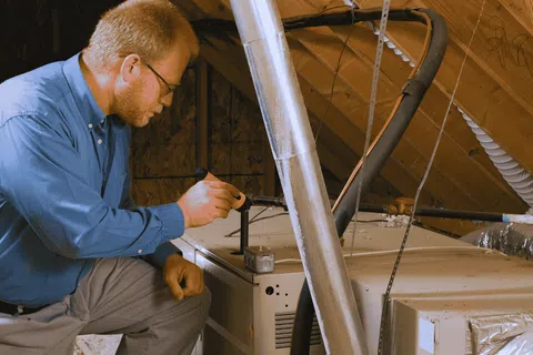 Technician inspecting and repairing HVAC system in an attic with tools and ductwork visible