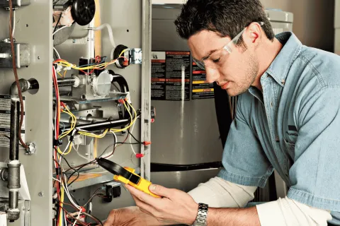 Technician wearing safety glasses inspecting furnace wiring with a multimeter in a mechanical room.