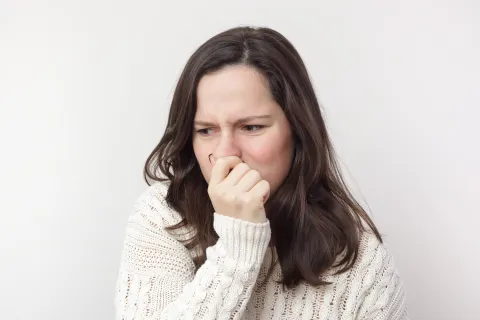 A woman in a white sweater grimaces and holds her nose, displaying a reaction to an unpleasant smell.