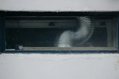 Small rectangular basement window with a ventilation duct visible through the glass in a white wall.