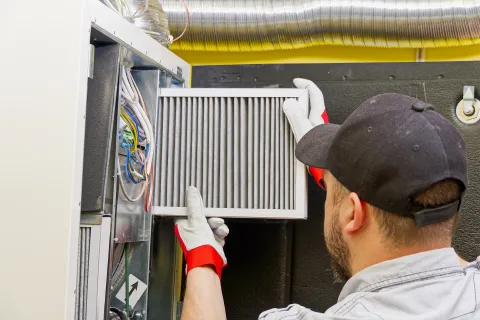 Technician replacing an air filter in a furnace with tools and gloves in a well-lit room.