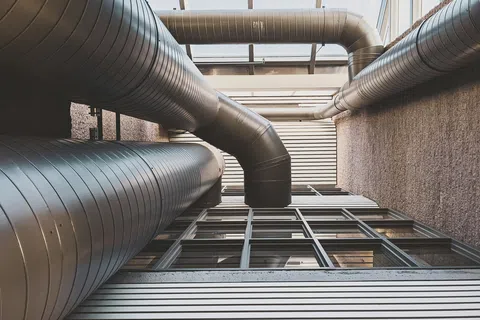 Industrial metal ventilation ducts above modern building windows and walls under a skylight ceiling.