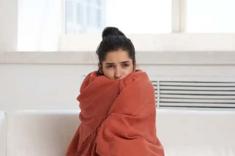 Young woman wrapped in a warm orange blanket sitting on a white couch looking cold and worried indoors.