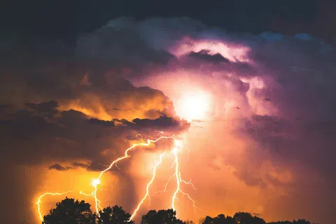 Multiple lightning bolts illuminating dark clouds over silhouetted trees at night with an orange and purple sky.