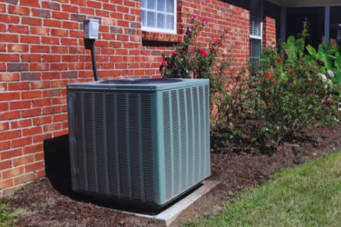 Outdoor air conditioning unit installed beside a brick house with a garden of flowering plants nearby.