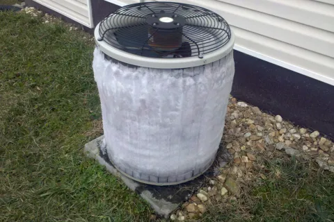 Outdoor air conditioning unit covered in ice and frost due to freezing temperatures next to house siding.