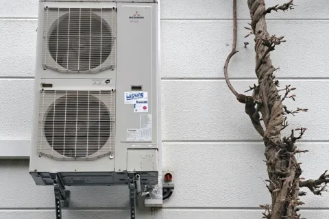 Outdoor air conditioning unit mounted on wall beside twisted leafless tree with climbing vine.