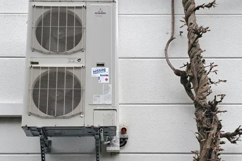 Wall-mounted air conditioning unit next to a bare twisted tree trunk against a white textured wall.