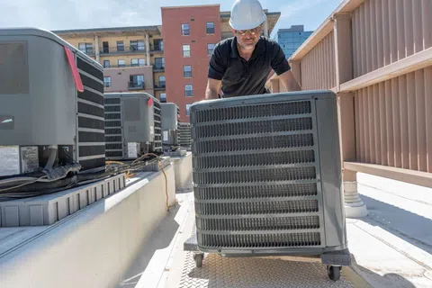 Technician wearing hard hat moves rooftop HVAC unit on wheels during maintenance at commercial building.