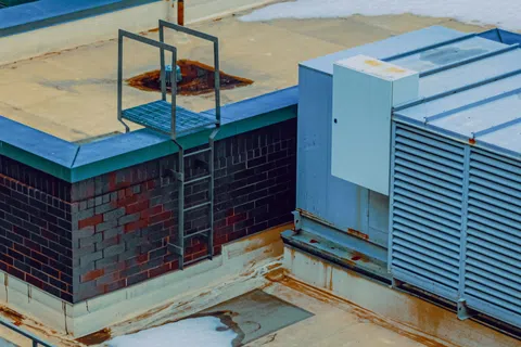 Industrial rooftop showing HVAC units, metal ladder, brick wall, and weathered concrete surfaces with some snow patches.