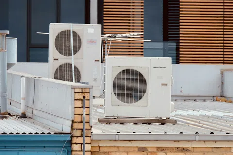 Air conditioning units installed on a rooftop against a building with wooden blinds on windows.