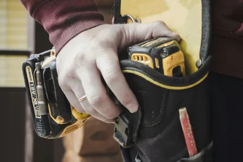 Close-up of a person wearing a tool belt holding a cordless drill near a wooden wall.