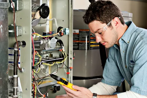 Technician wearing safety glasses inspecting and repairing electrical wiring inside a home furnace unit.