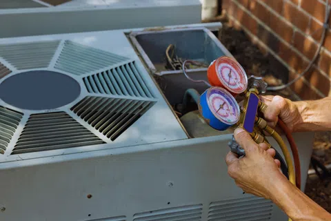 Technician using pressure gauges to check air conditioning unit maintenance outdoors near brick wall.