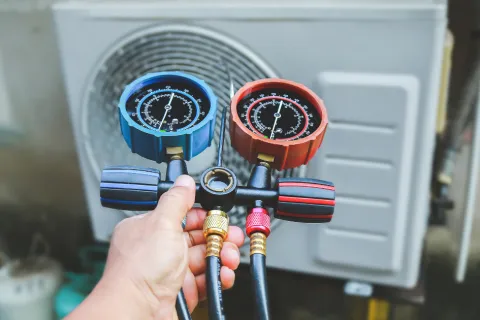 Hand holding HVAC manifold gauge set with blue and red dials in front of an air conditioning unit outdoor.