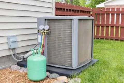 Residential air conditioning unit with refrigerant tank and pressure gauges installed outside house near grass and fence.