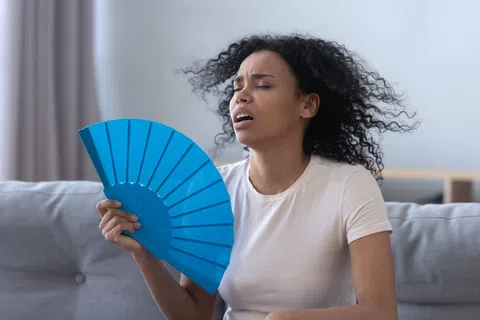 Woman in a white shirt fanning herself with a blue hand fan, appearing hot and uncomfortable indoors.