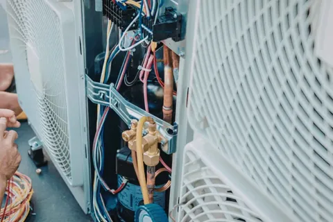 Technician repairing and wiring components inside an outdoor air conditioning unit with visible copper pipes and wires.