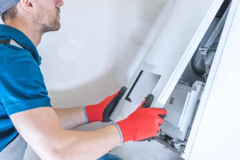 Technician wearing red gloves repairing or installing a white boiler or HVAC unit on a wall indoors.