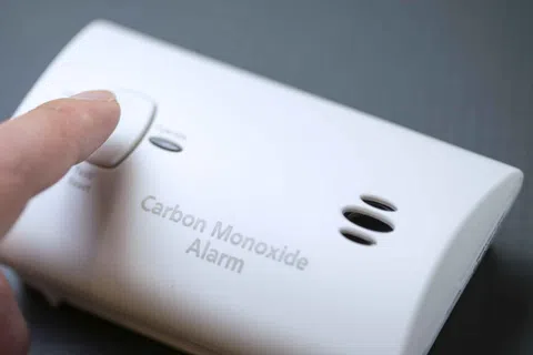 Finger pressing test button on white carbon monoxide alarm mounted on a wall.