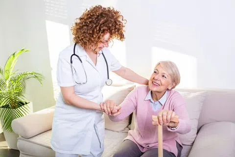 Nurse with stethoscope helping elderly woman with cane sitting on couch in bright living room