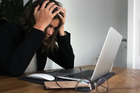 Stressed woman holding head sitting at wooden desk with laptop, phone, glasses, and mouse, working indoors.