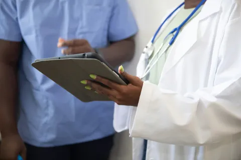Doctor in white coat holding a tablet with a nurse in blue scrubs standing beside them, discussing patient care.