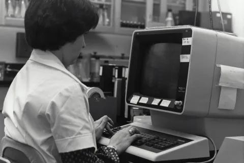 Woman in a lab coat working on a vintage computer showcasing aging IT technology in healthcare.