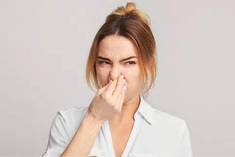 Woman in white shirt holding nose with a disgusted expression against a neutral background.